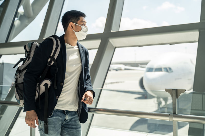 Homem de negócios viajante asiático usando máscara facial esperando para embarcar no avião, parado no terminal de partida no aeroporto.